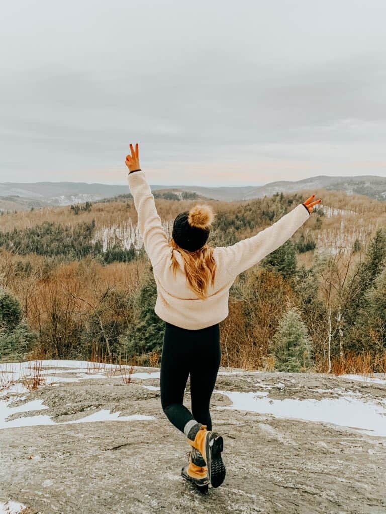 Girl standing with her arms up on a rock covered in snow looking out to the mountain landscape while hiking near Burlington, Vermont.
