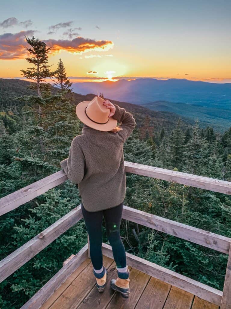 Girl holding her hat looking out to the sun setting over blue mountains on a hike near Burlington VT.