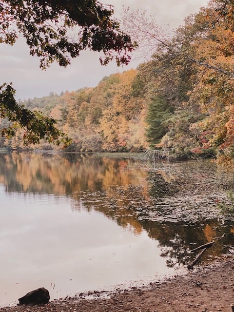 A scenic lake is seen with autumn foliage surrounding and reflecting in the waters under a cloudy sky