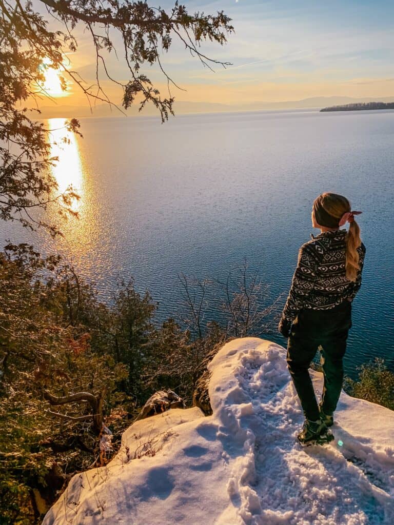 Girl standing on a snowy rock edge looking out to the water on hiking trails near Burlington, Vermont.