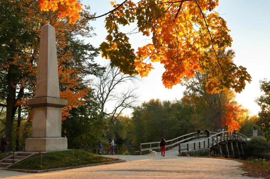 People walking on a bridge surrounded by trees with fall colors under a blue sky in Concord, Massachusetts.