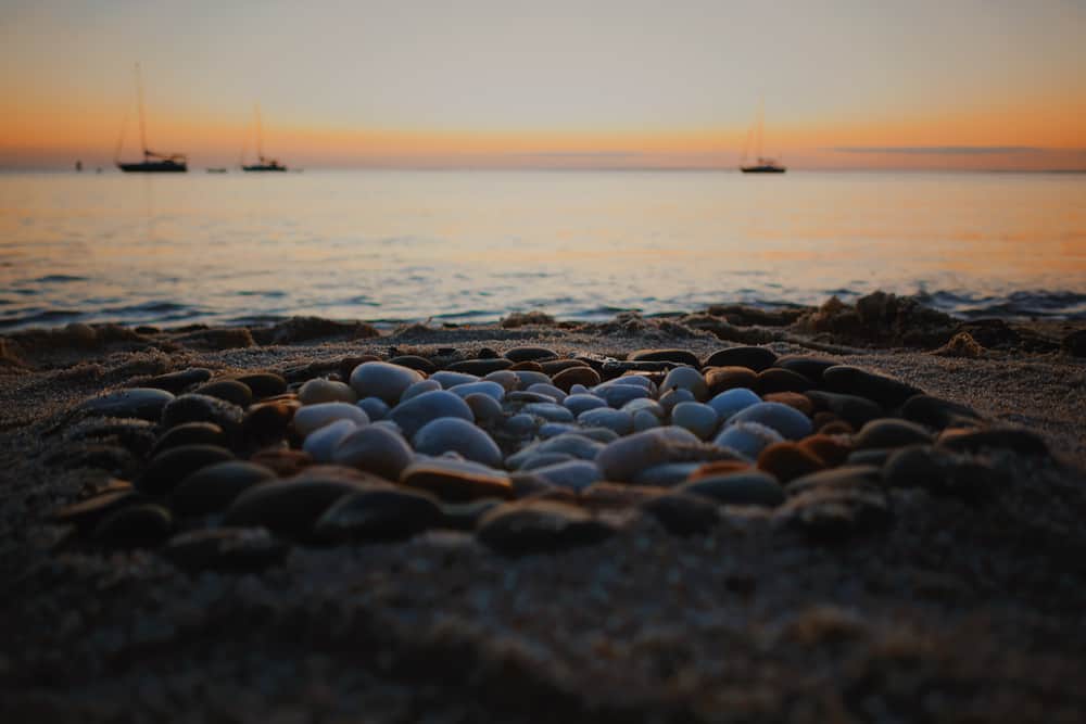 A pile of rocks is seen close up on a sand beach with a calm sea beyond under a softly setting sun in the distance.