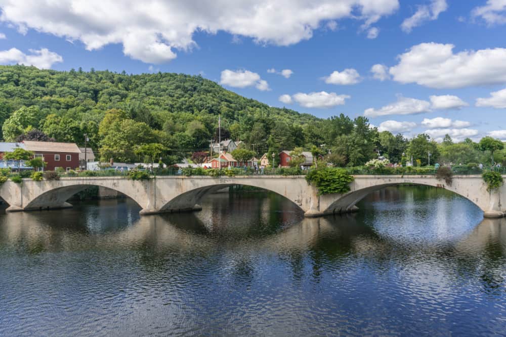 A bridge made of stone stretches over a body of water. Behind the bridge is a small hill covered in forestry, under a blue cloudy sky in Western Massachusetts