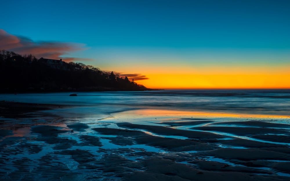 A clear blue water beach is seen under a blue and orange sky during sunset.