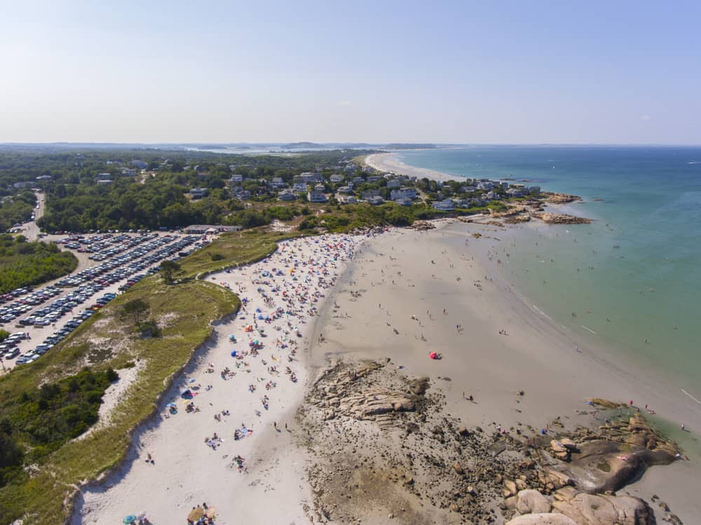 A beach is pictured from above with soft sand filled with beachgoers and clear calm water stretching out for miles