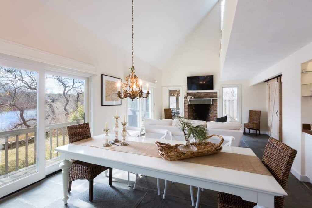 Interior of a white walled home. A long white dining table is in the foreground surrounded by white and brown wicker chairs. A historic chandelier is above the table. In the distance is the living room with a white plush couch facing a television mounted over the top of a brick fireplace.