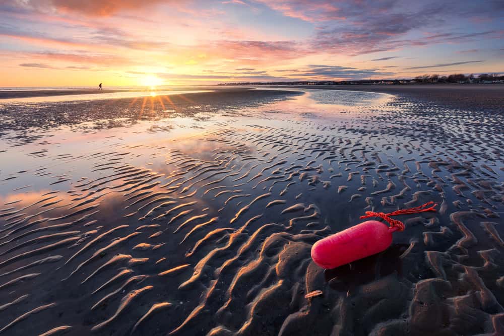 A sun sets over a sandy coastline, where a red buoy lays at low tide.