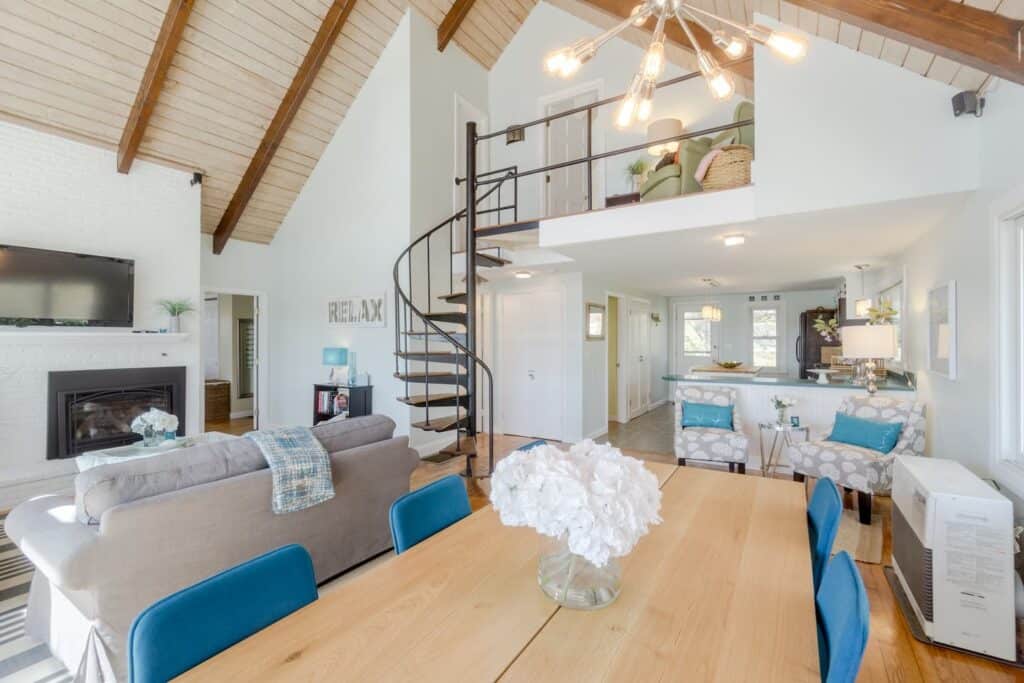 Inside of a home with two floors. In the center, a light wooden table is surrounded by turquoise chairs. To the left, a grey couch can be seen. The background has a kitchen. The second floor has a sitting area, lofted.