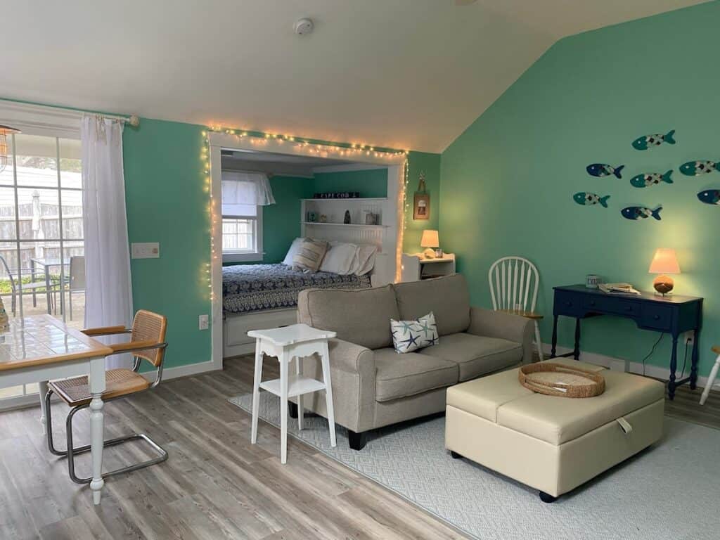 Interior of a rental home with turquoise walls. The main area features a grey couch by a tan table. Through the back door behind the couch is a bed.