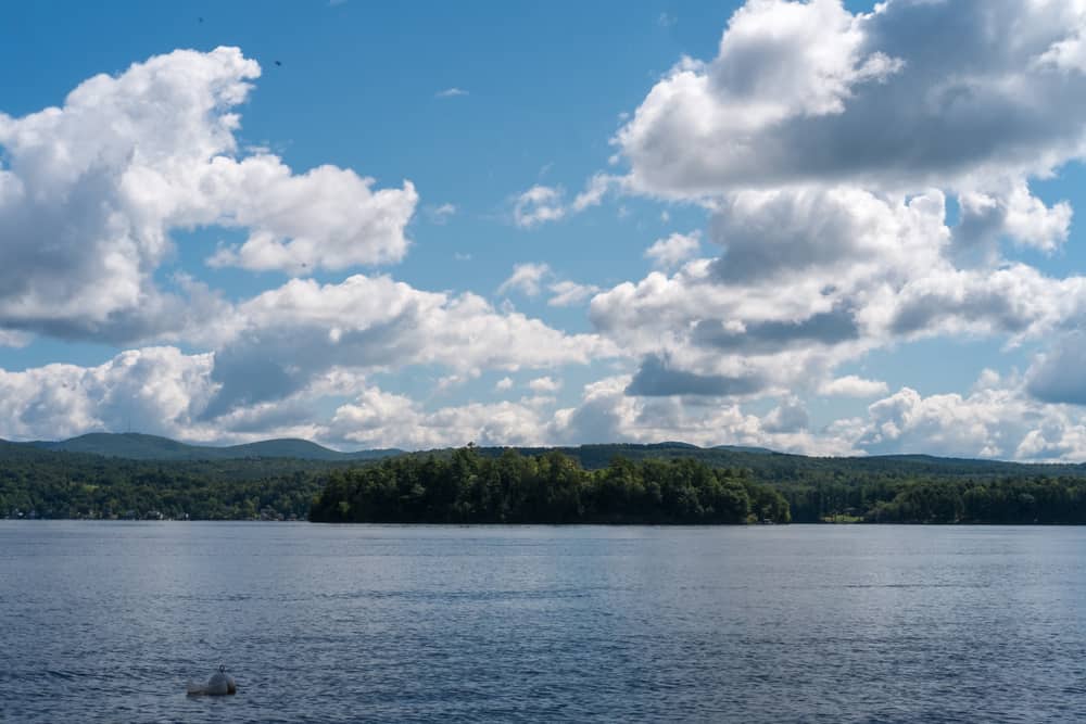 Calm view of a lake under a blue sky dotted with clouds. In the distance, on the other side of the lake, is a forest.