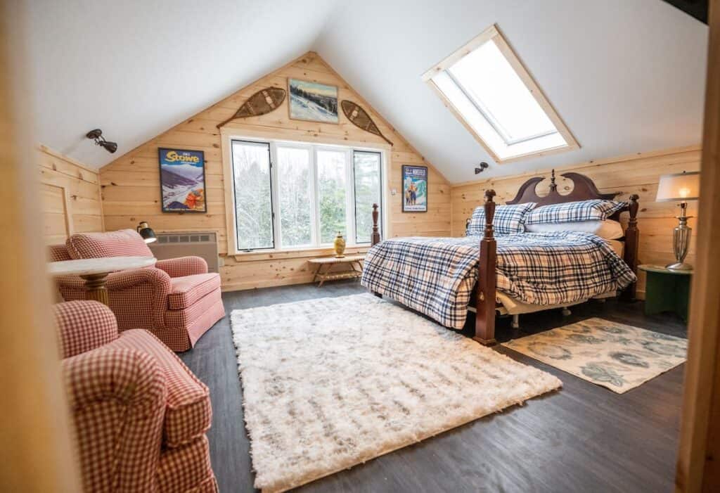 Interior of a bedroom with warm wooden walls. A bed is made with plaid bedding. Snowshoes and other winter memorabilia hang from the walls. A skylight is situated above the bed, letting in warm lighting from outdoors.