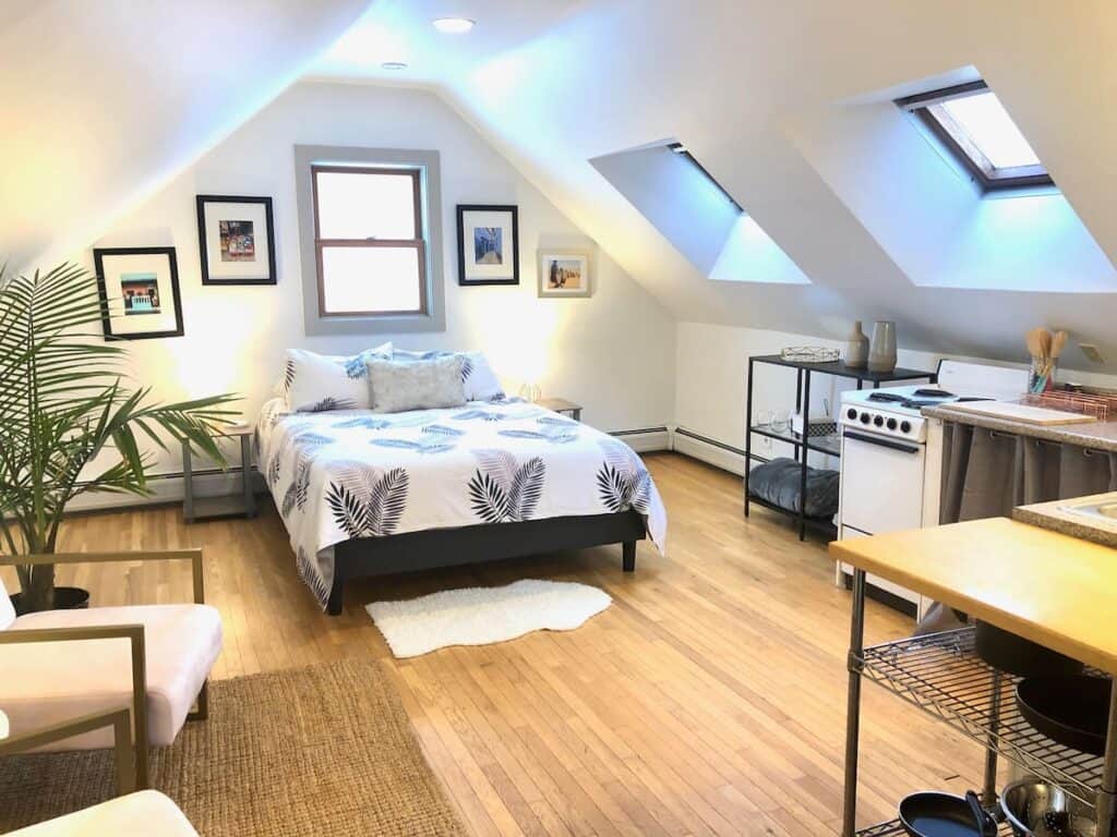 Bedroom with white walls and an A-frame ceiling, featuring two skylights. A bed is made with black and white bedding, featuring plants. To the right of the bed is a small kitchen area with a stove and oven.