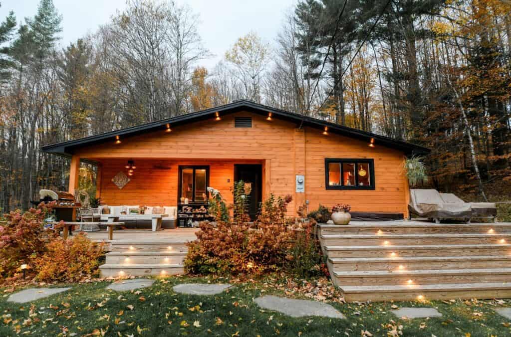 Wooden cabin with black window frames and a black door. Stairs lead up to the home, made out of wood, featuring lighting. The home is surrounded by a forest.