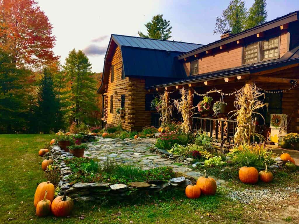 Exterior of a wooden log cabin in fall surrounded by trees. Pumpkins are situated around the front of the home.