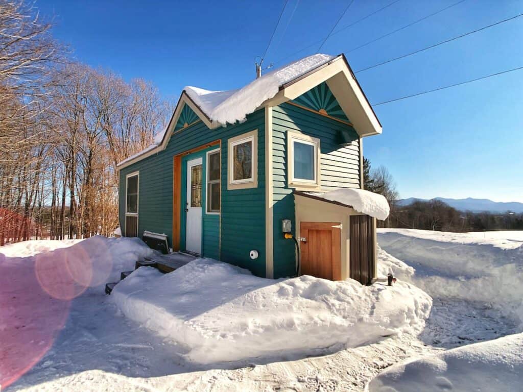 Teal tiny home covered in snow under a blue sky.