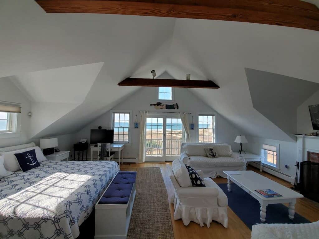 View of the inside of a bedroom. A bed is made on the left with a white and blue comforter. Two white couches are situated around a white table on a blue rug on the right. The background has large doors that offer a view of the beach.