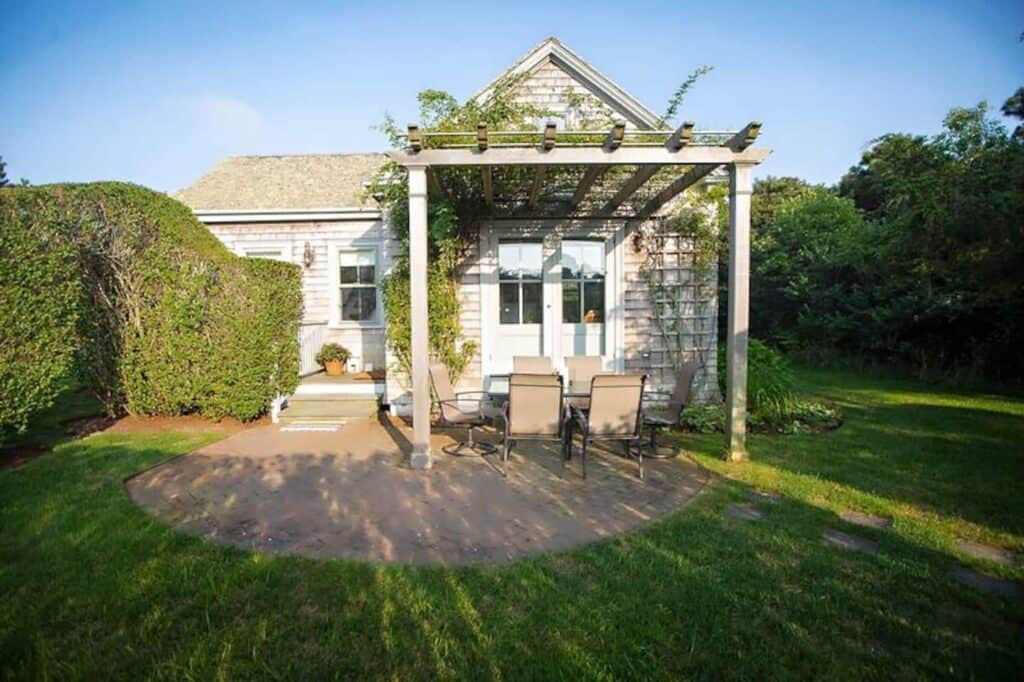 Exterior of a cottage with a table and chairs set up outdoors. Greenery grows on the side of the house.