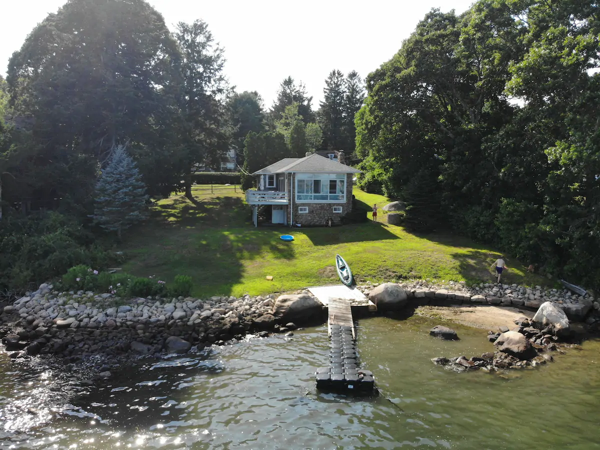 Quaint neutral-colored cottage on a hill by a lake. A dock leads out to the water.