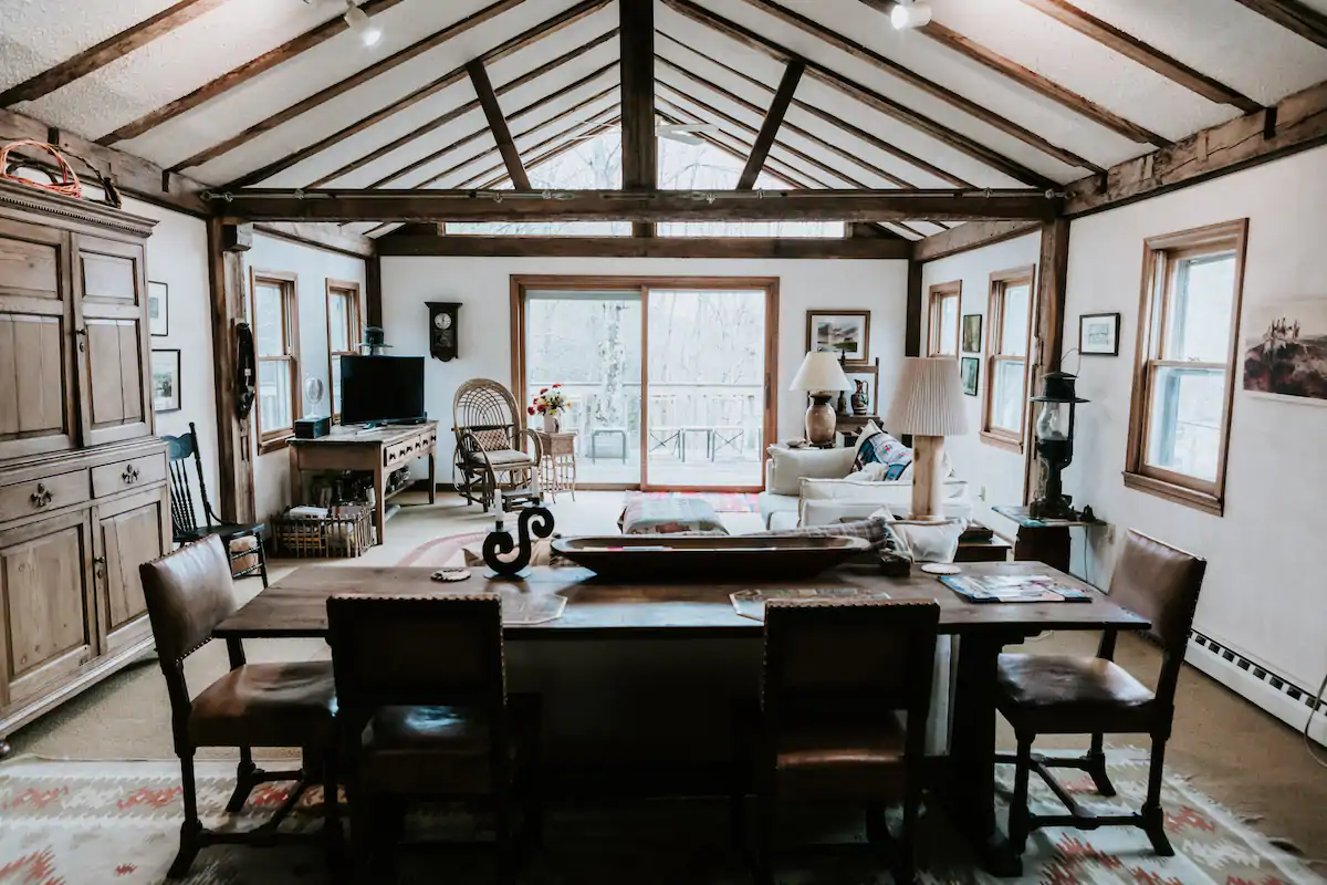 Interior of a dining and living room. The ceiling is white with dark wood exposed beams. In the foreground, a dark wood dining room table is sitting surrounded by chairs. In the background, there\'s a living room setup with a cream couch and a TV on a desk.