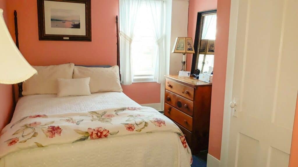 Interior of a bedroom with pink walls. A bed is made with white bedding and a blanket with a floral pattern is folded on top of it.