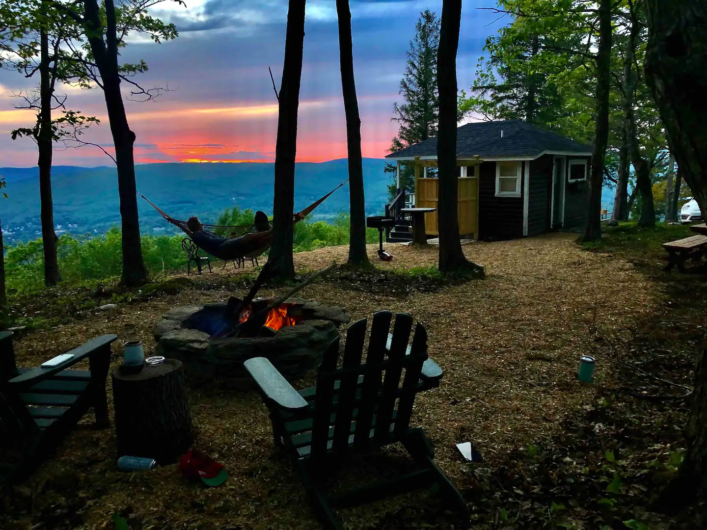 In the distance, there\'s a sunset sky with orange and hues of purple. In the foreground, chairs are set up around a crackling fire. A person is relaxing in a hammock by a small brown cabin.