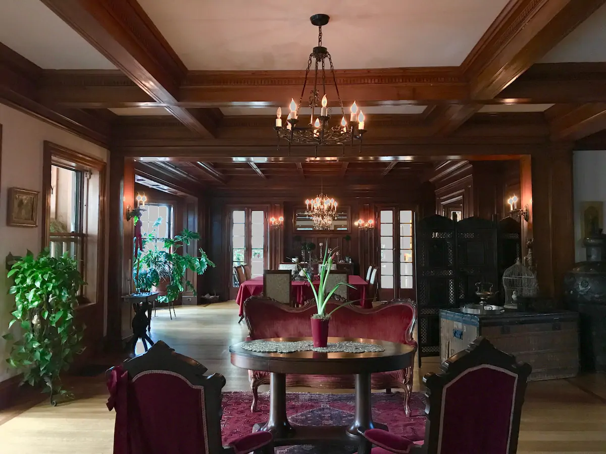 Inside of a historic home with wooden beams. A chandelier hangs from the ceiling above two red velvet chairs next to a table. In the background is another dining room set.