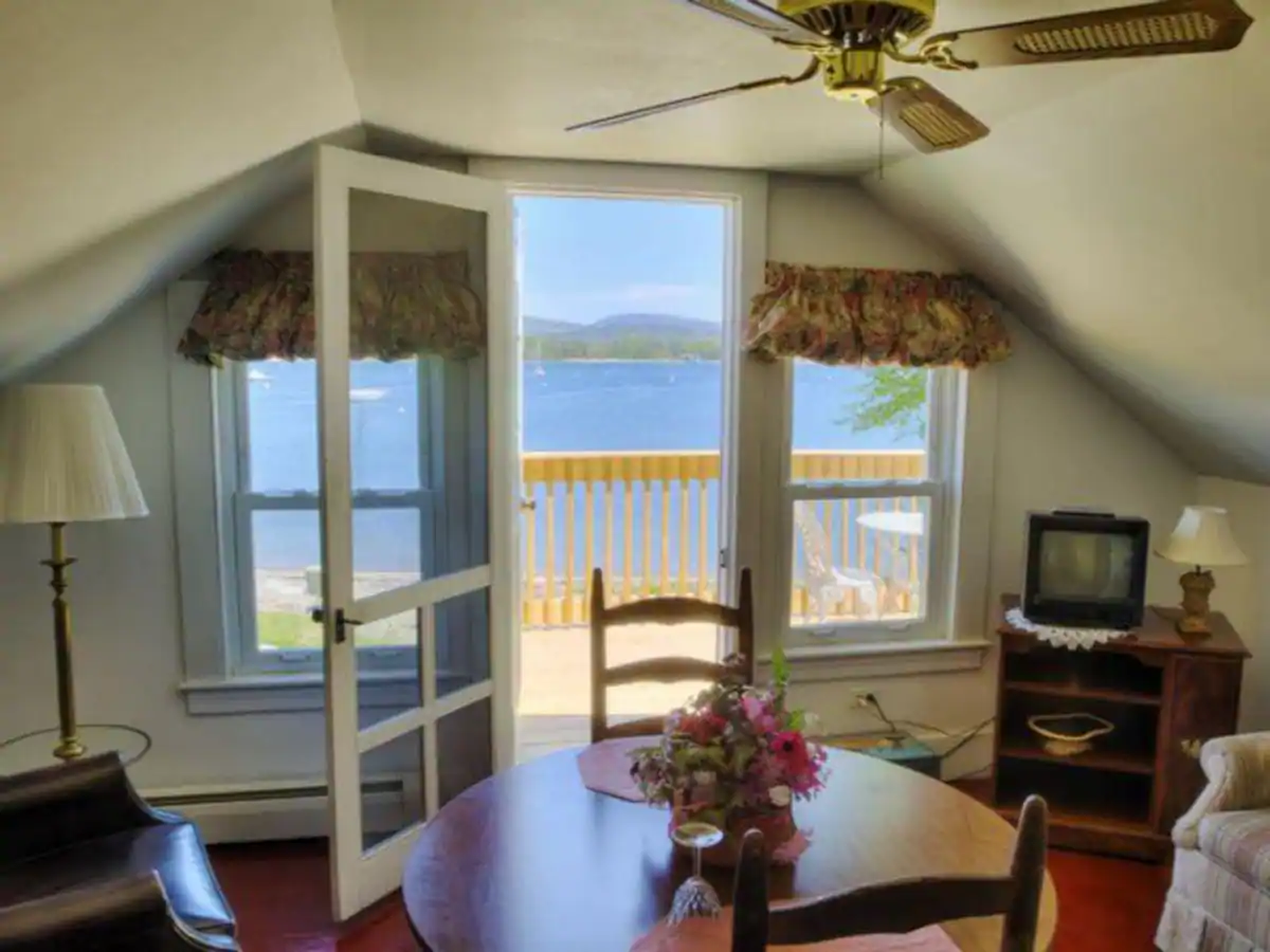 Interior of a home with white walls. Two chairs sit around a circular dining table. A door leads out to a view of a blue lake surrounded by mountains.
