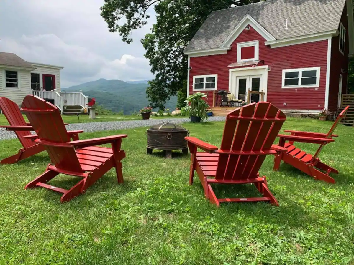 Red chairs set up next to a fire pit in the grass. In the distance, there\'s a red home and rolling mountains.
