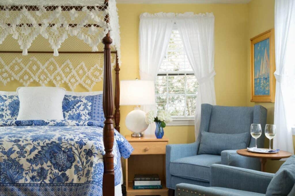 Interior of a yellow room. A bed with blue and white bedding is made on the left. A side table is next to the bed, and a blue plush chair is seated to the right of the side table.