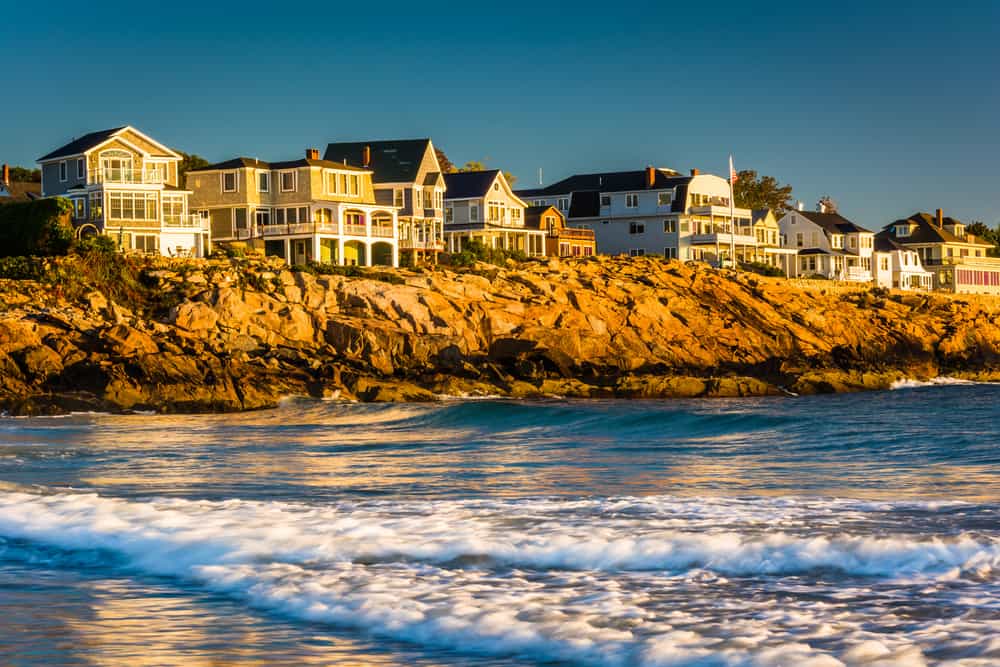 Historic homes set against a rocky cliff under a blue sky. Ocean waves crash up against the sandy beach in Maine.