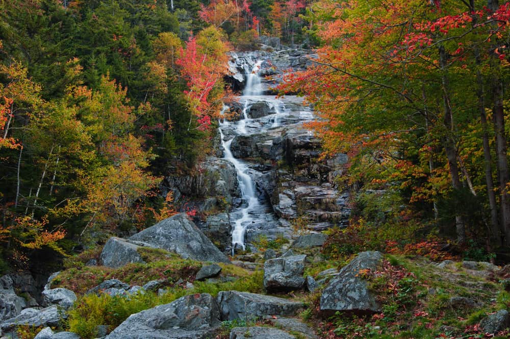 A waterfall trickles down a series of rocky edges surrounded by trees with red, yellow, and green leaves.