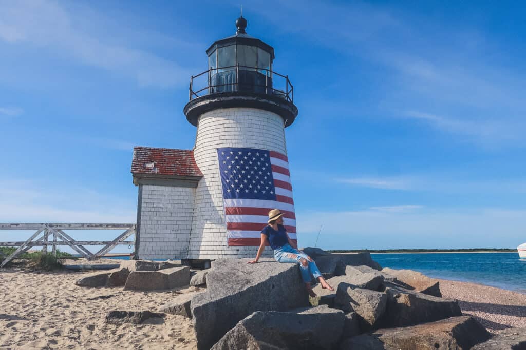A white lighthouse with an American flag painted on it sits under a deep blue sky; jagged rocks are beside the lighthouse and a woman is perched on top of them.
