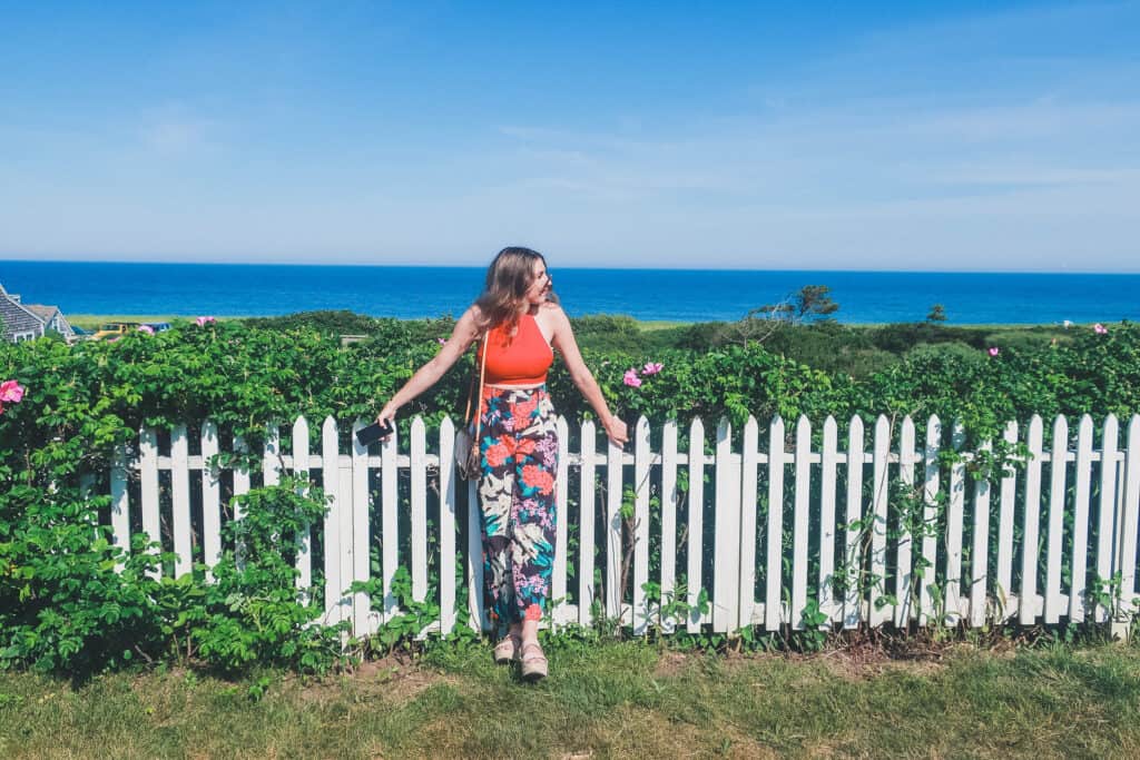 A woman is leaning against a white picket fence that is overgrown with pink roses; the deep blue ocean and clear blue sky is seen in the distance behind her