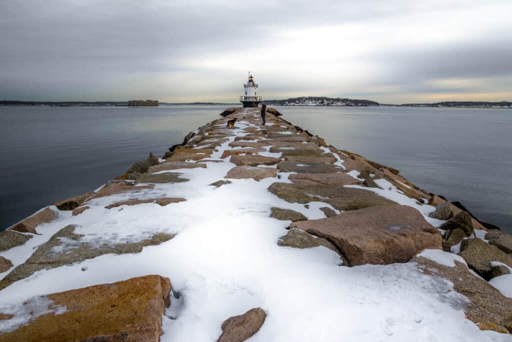 Stone walkway covered in snow leading to a Portland Maine lighthouse in the distance by the water under a grey sky.