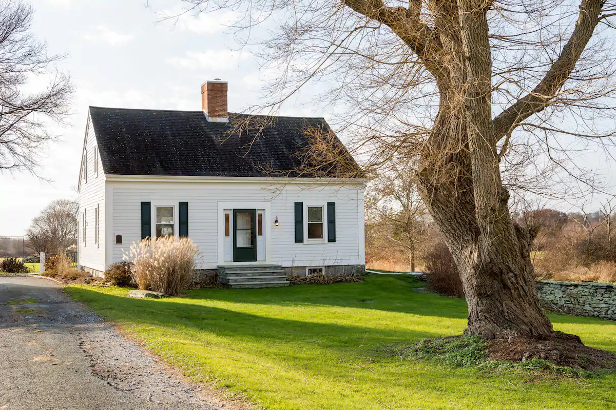 White farmhouse sits on a plot of land by a dead tree under a blue sky.