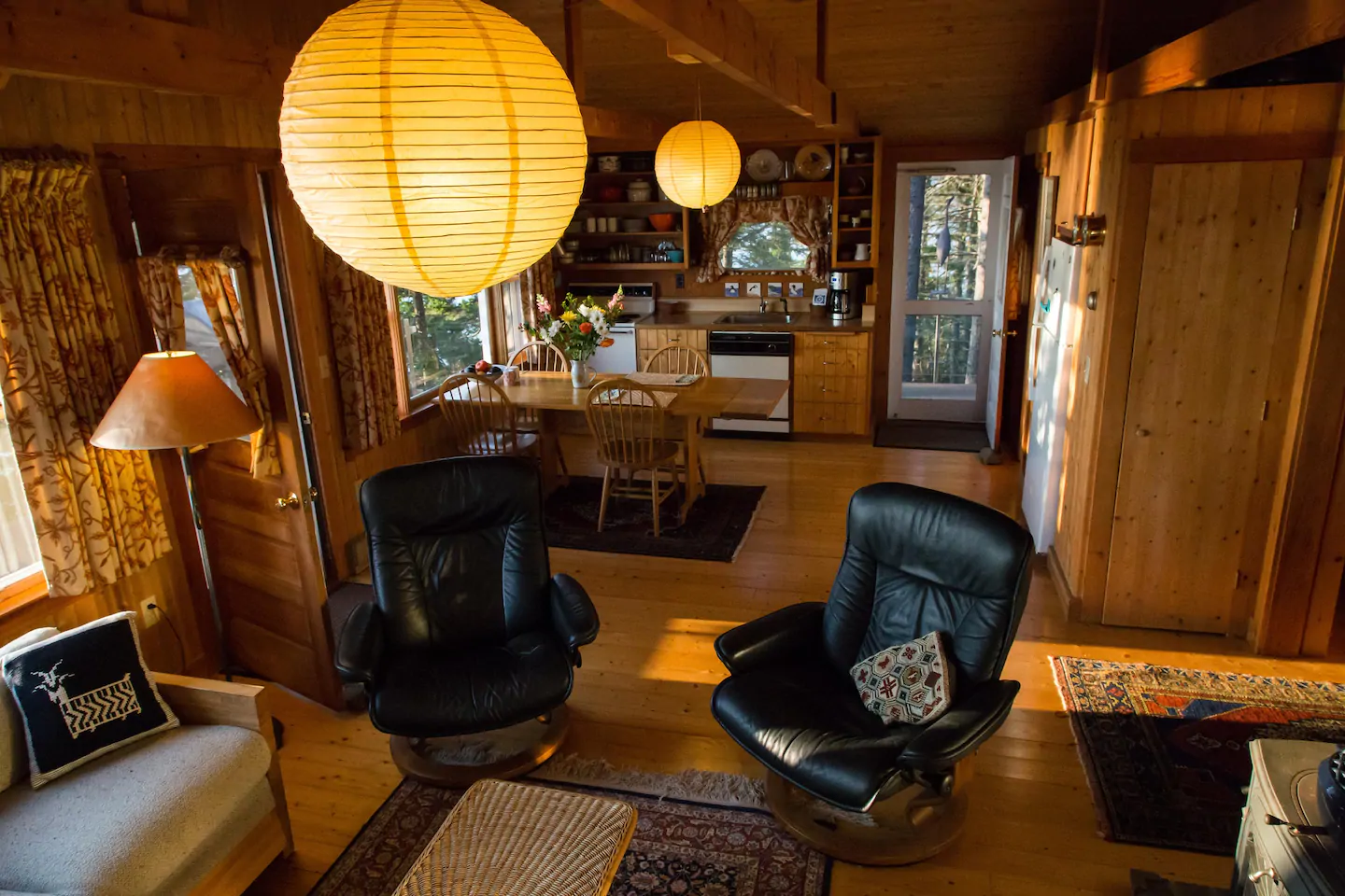 Interior of a log cabin with warm lighting. The foreground features two leather chairs and a coffee table. The background has the kitchen with a wooden table featuring four chairs.