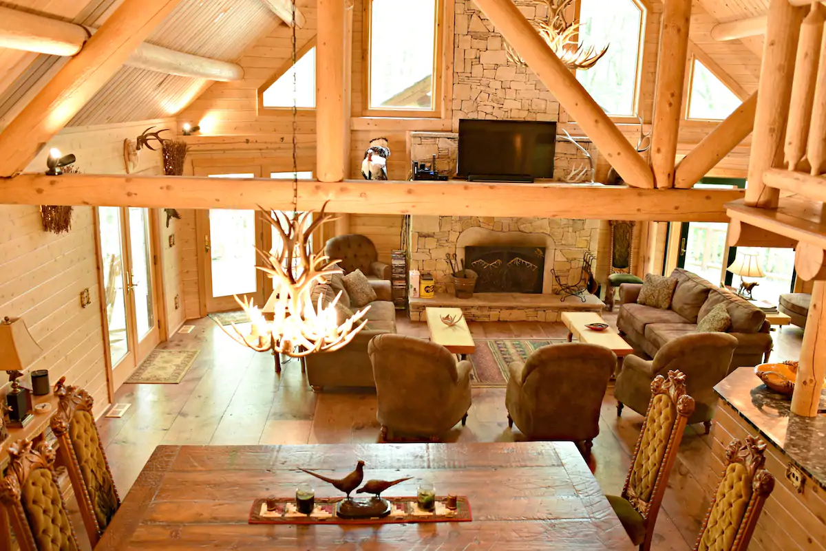 Interior of a lodge living room. Exposed wooden beams are on the ceiling and a set of comfortable chairs and couches surround a fireplace in the background. The foreground has a wooden table with chairs.