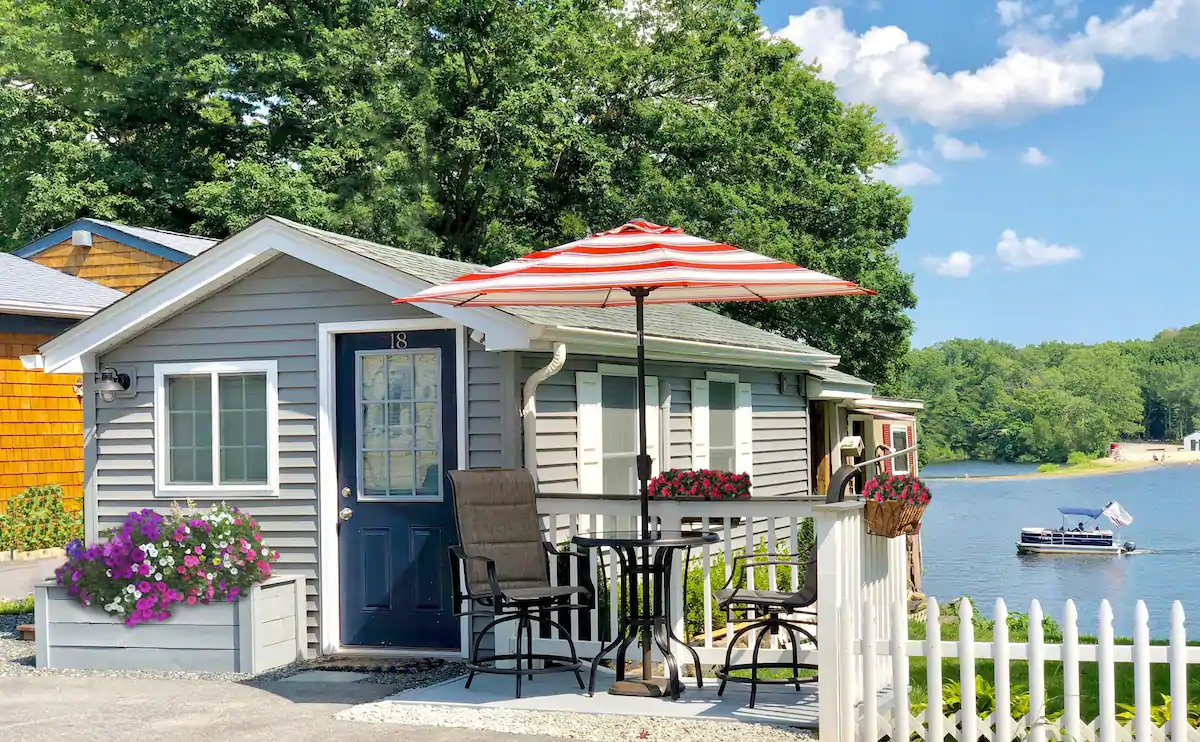 Grey home on the coast with a navy door. A table and chair set under a red and white striped umbrella is next to the home.