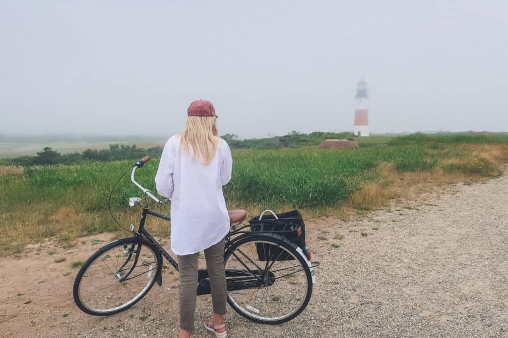 Woman with blonde hair standing next to a bike on a sandy beach. In the distance, a lighthouse can be seen under fog.