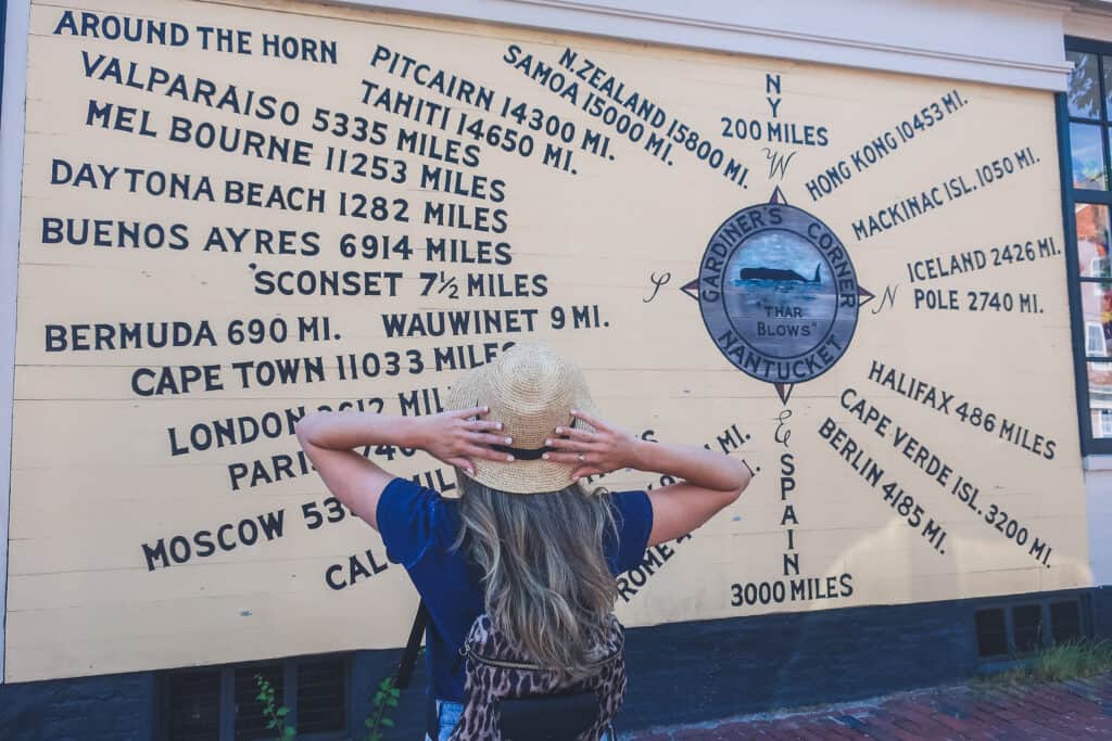 Woman holding her hat down while looking at street art that features distances to locations all around the world from where she is standing.