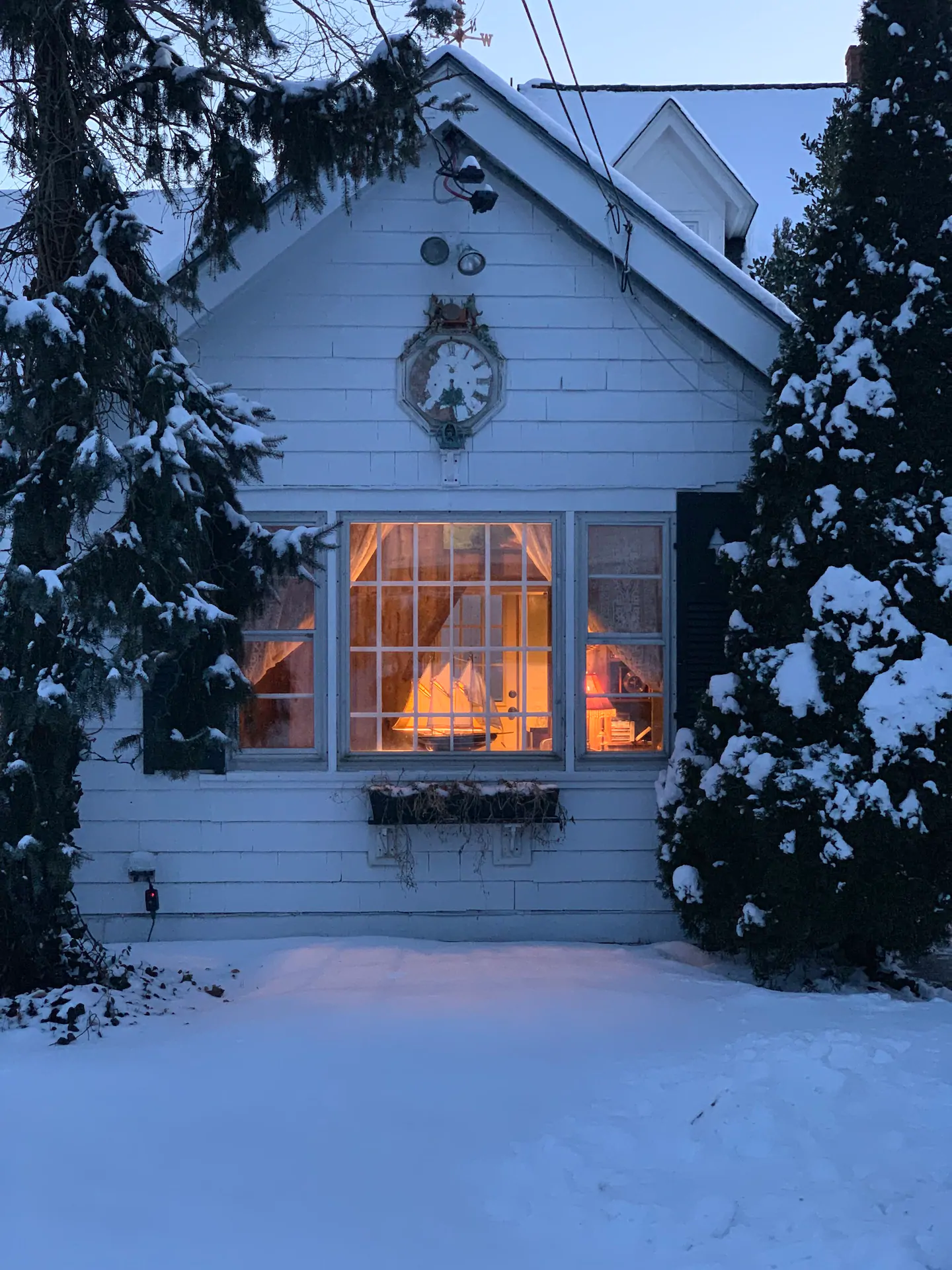 White home with large windows. Warm lighting can be seen through the windows. Snow covers the exterior of the home, as well as the trees around it.