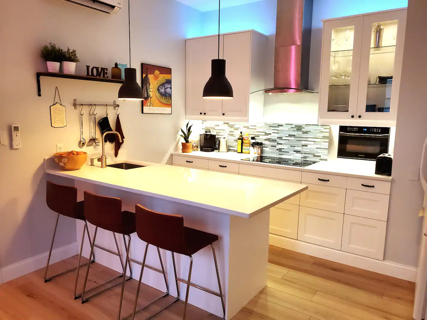 Interior of a kitchen. Three brown chairs are sat up against a white island, and kitchen utensils hang from the walls. 