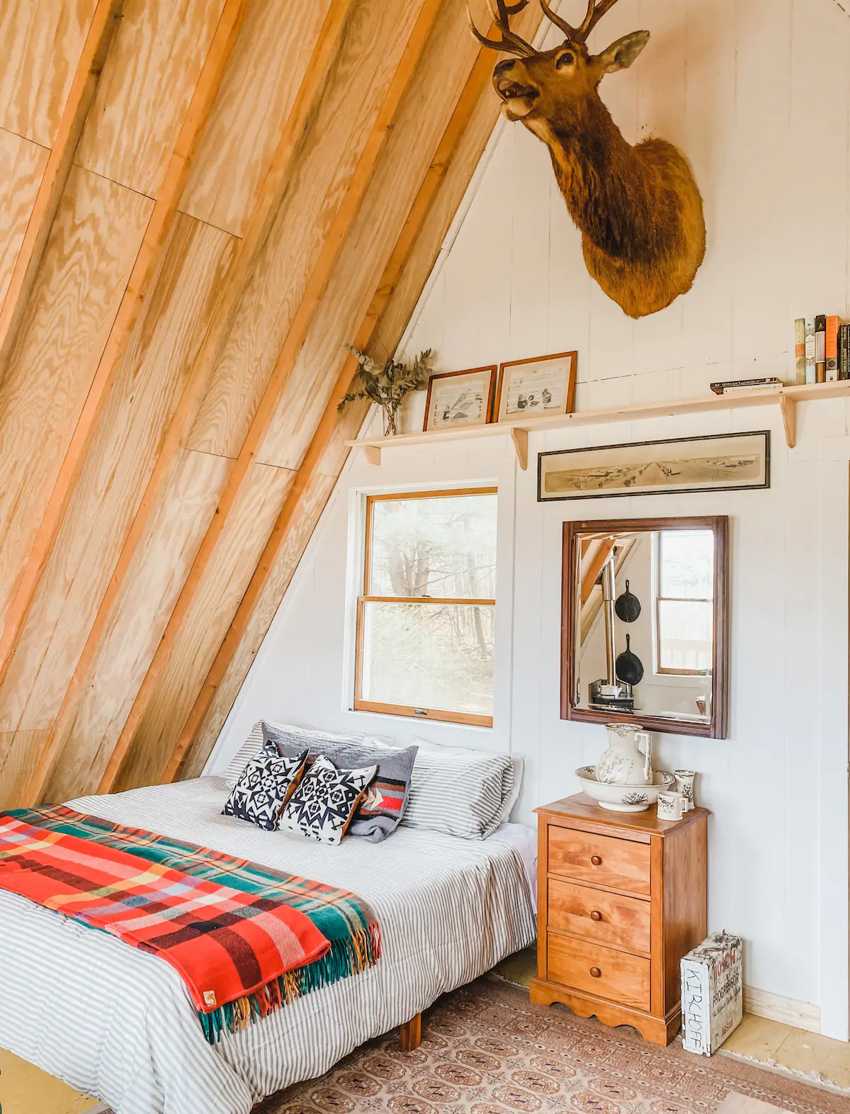 Bedroom with a slanted wooden ceiling. The bed has a red and green plaid blanket on it and a window is above the bed frame.
