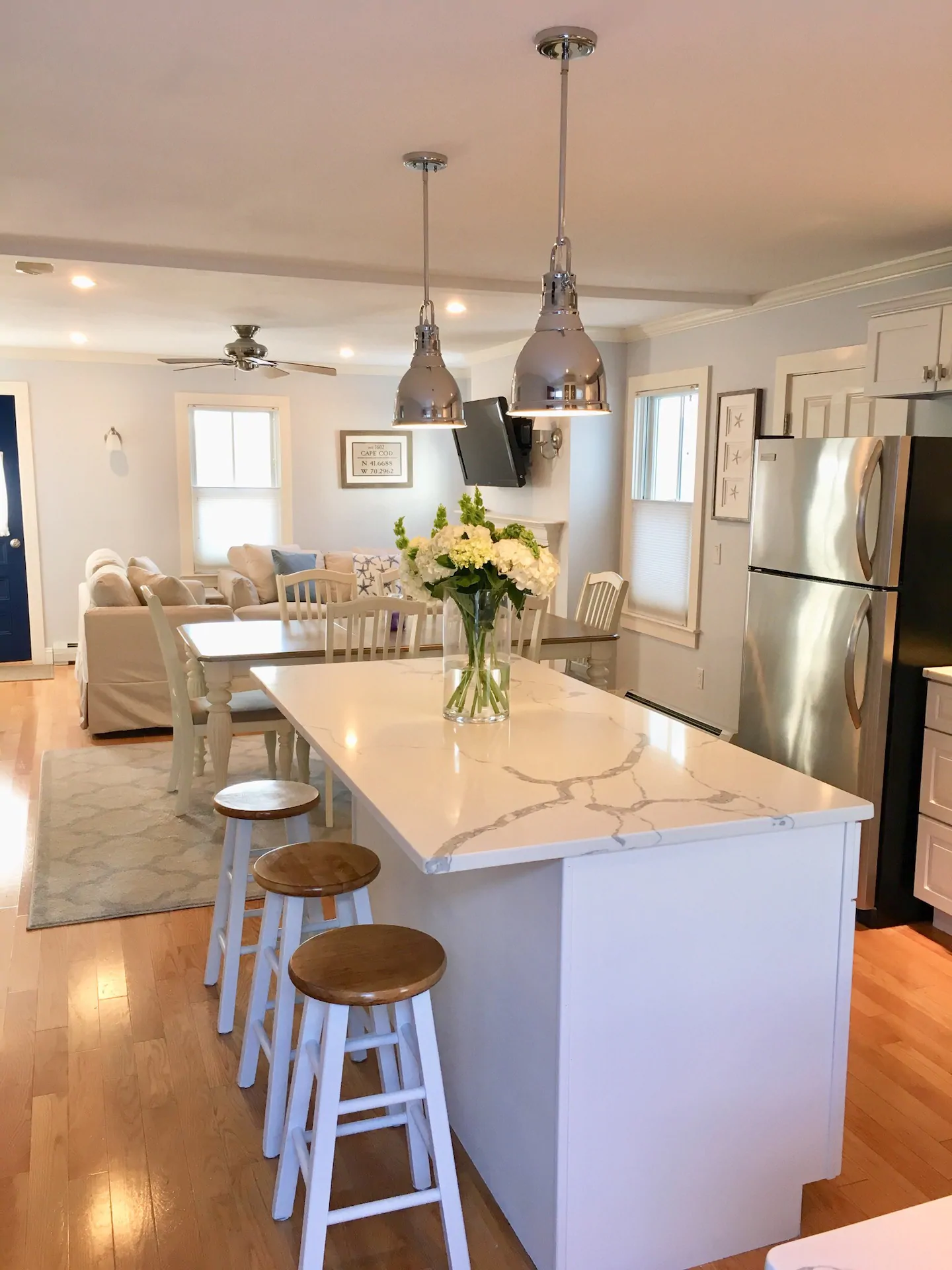 Kitchen interior with three stools set up against a white marble kitchen island. A living room with a couch and a fan can be seen in the background.