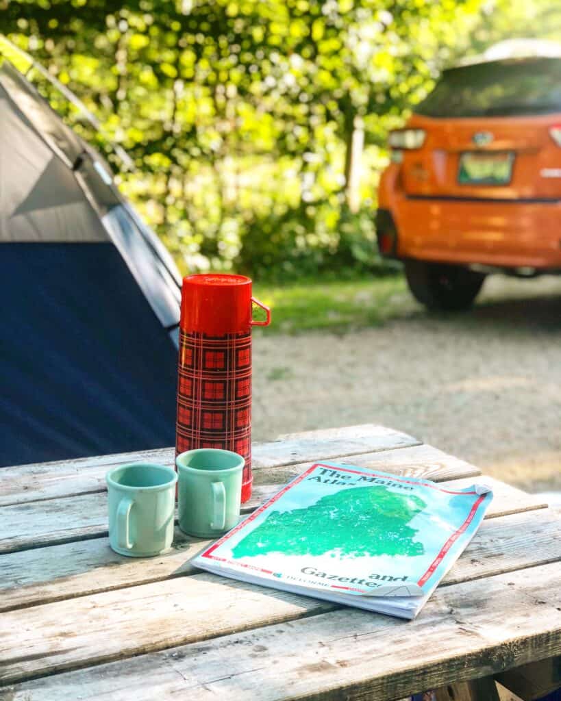A close up of a picnic table with a red thermos and 2 cups, and the back of an orange car to the right
