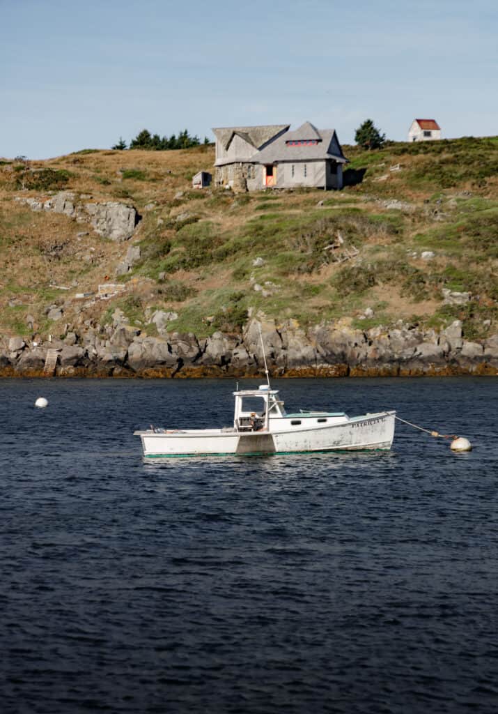 In the foreground, a white boat floats on the water. In the distance, a grey home sits on a cliff.