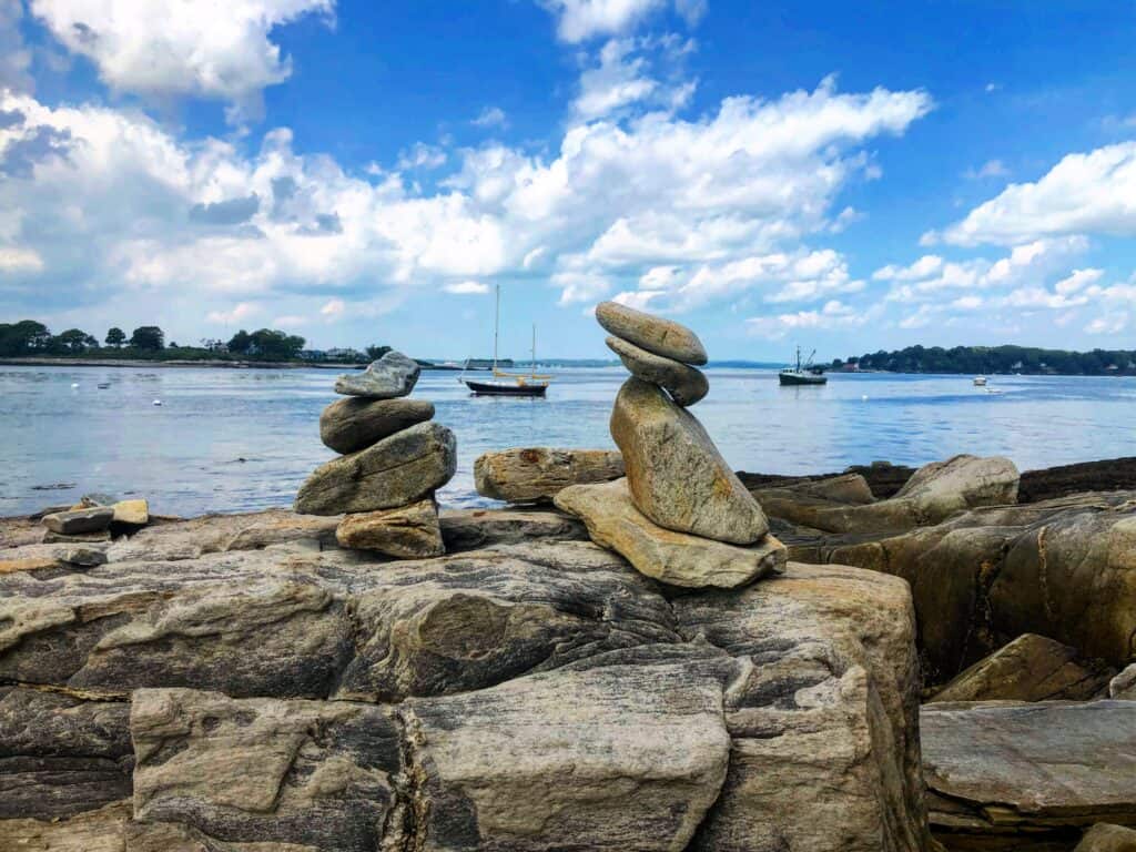 Rocks stacked on top of each other by the water. Boats float on the water in the distance.