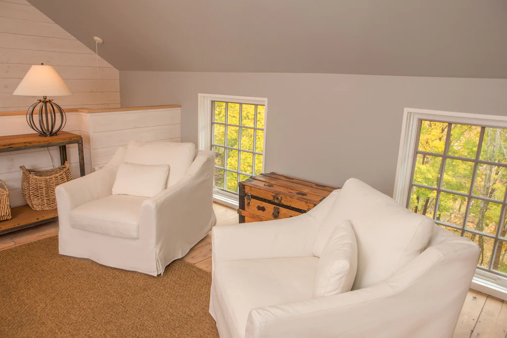 Two white living room chairs with pillows against a grey wall with 2 windows