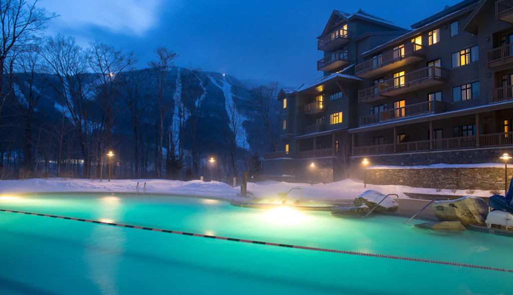 A swimming pool in front of the snowy ground and a chalet style hotel