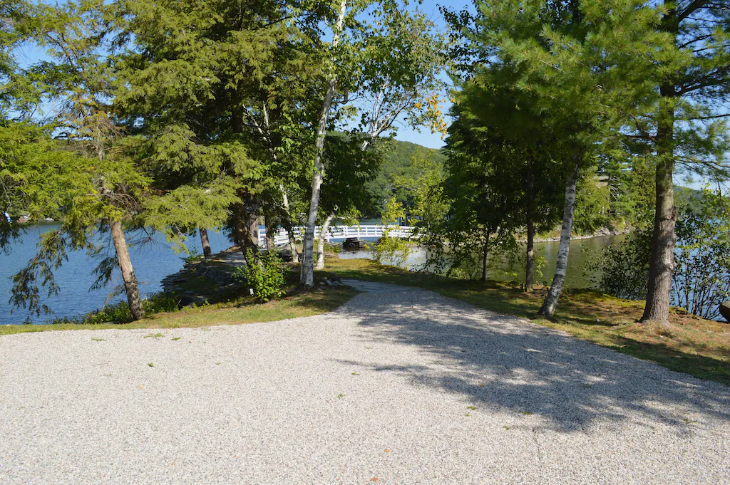 A wide gravel driveway can be seen leading to a white footbridge surrounded by a tree-lined lake on a sunny day